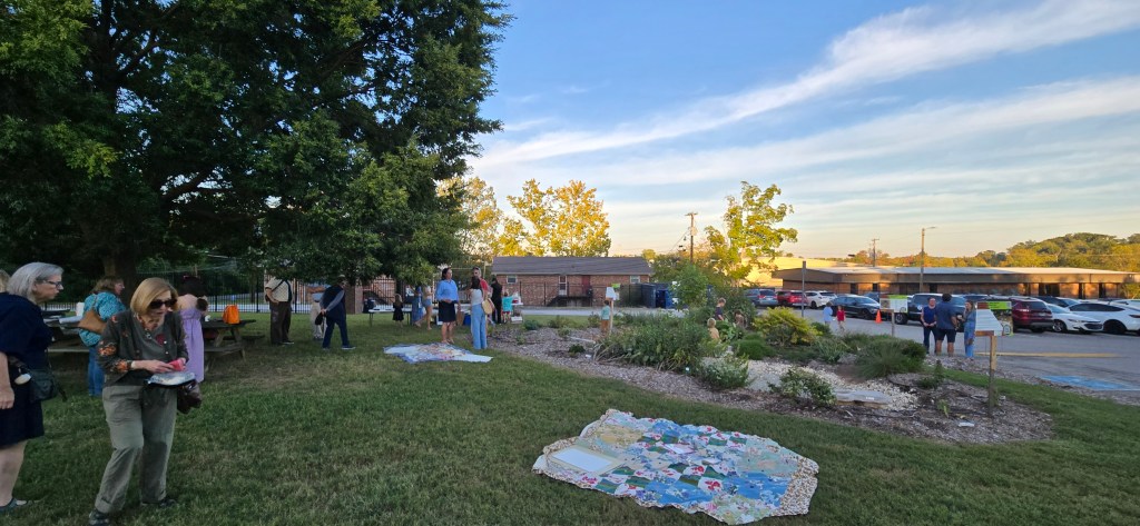 A community gathering in a park featuring several people interacting, with a picnic area and a garden in the background. The scene shows a clear sky and trees.