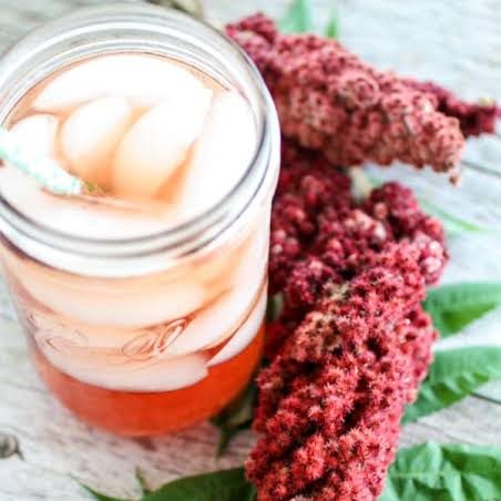 A refreshing drink in a mason jar filled with ice, accompanied by clusters of red sumac flowers and green leaves on a wooden surface.